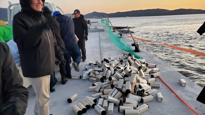 People standing next to a pile of bottles with rice, American one dollar bill, and a Bible on a flash drive, on a boat on the Yellow Sea. 