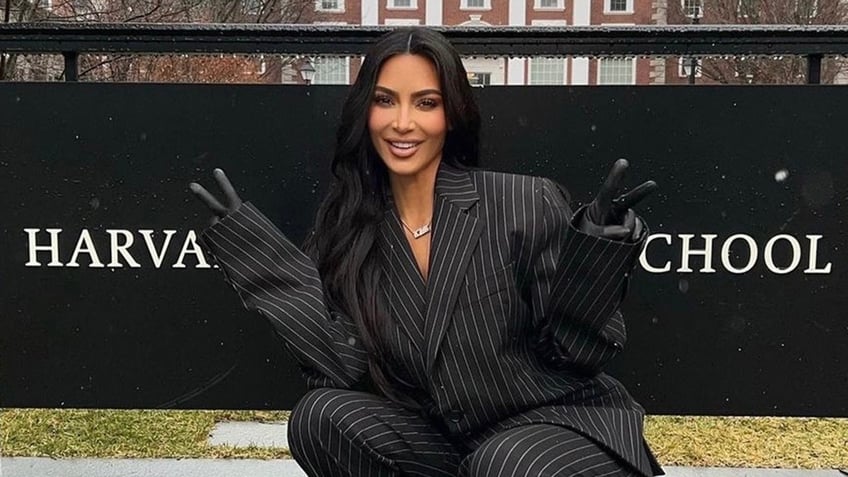 Kim Kardashian in a black and white pinstripe suit gives to peace signs in front of a sign that says 'Harvard Business School'