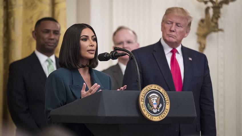 Kim Kardashian talks with her hands behind the podium at the White House with former President Trump looking on