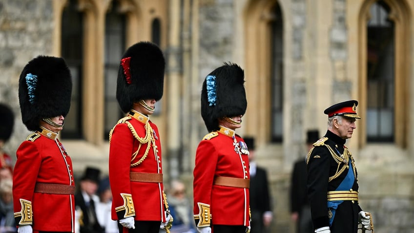 King Charles standing in front of British guards in uniform.