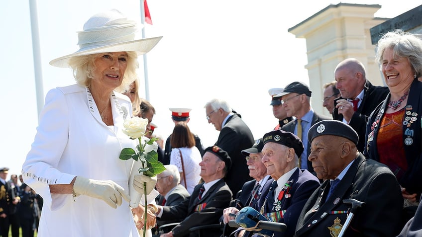 Queen Camilla wearing all white holding a rose as she walks next to a row of veterans sitting down in uniform and smiling.