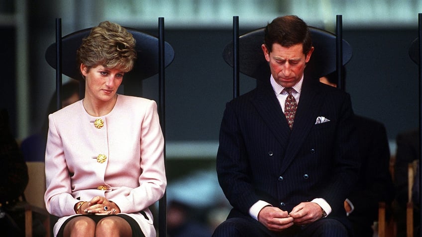 Prince Charles looking down as Princess Diana looks away as they sit down next to each other.