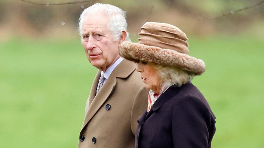 King Charles in a tan coat walks alongside wife Queen Camilla in a dark coat and tan hat