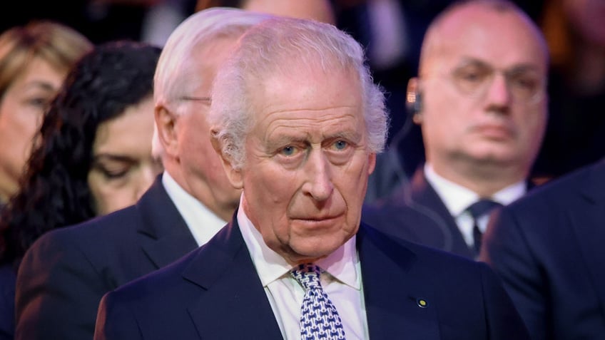 A close-up of King Charles looking serious in a navy blazer, white collared shirt and a multiprinted tie.