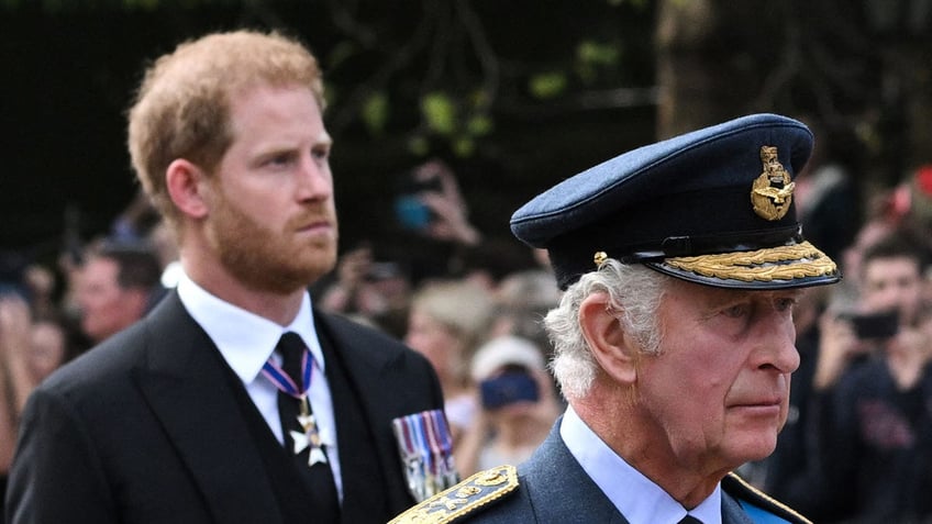 Prince Harry in a suit walking behind his father who is in uniform looking somber