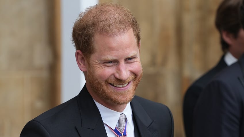 Prince Harry smiles in a black suit and white button down with medals adorned on his suit at the coronation of King Charles