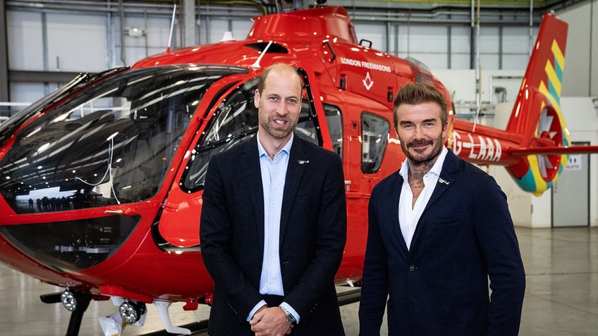 Prince William standing next to David Beckham in front of a red helicopter.