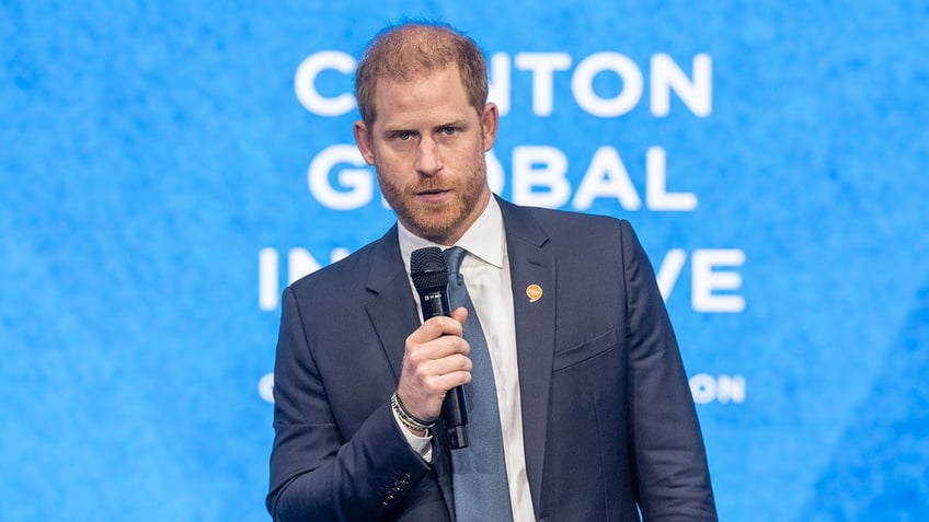 Prince Harry wearing a blue blazer speaking to a mic in front of a light blue sign.