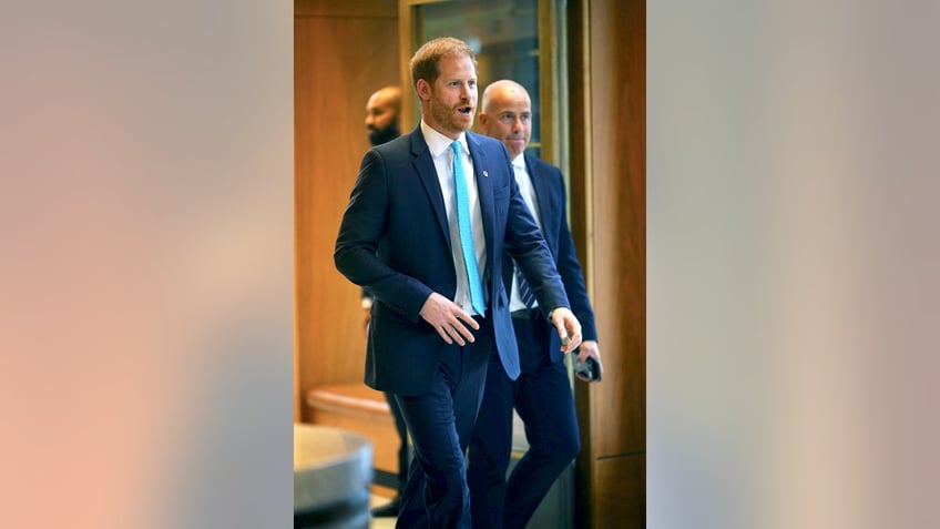 Prince Harry in a navy suit walking ahead of two men in matching blue suits.