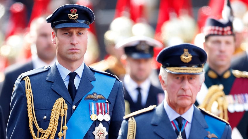 Prince William and King Charles in uniform walking together and looking somber.