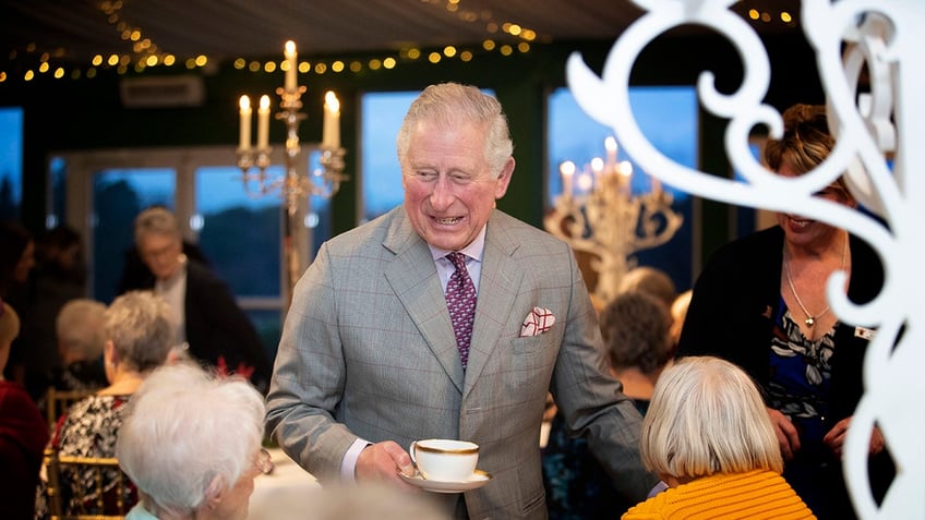 King Charles holding a teacup and smiling in a grey suit.