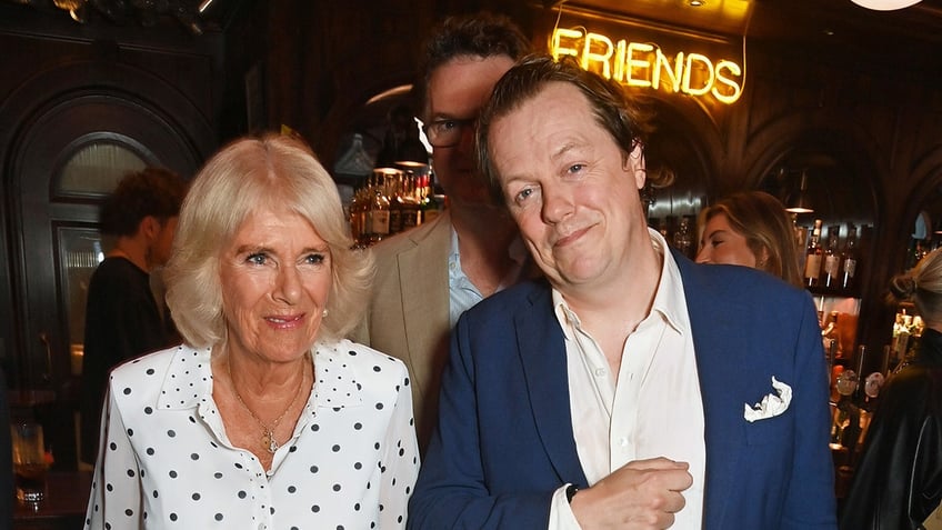 Tom Parker Bowles with a blue blazer and white shirt holding the arm of his mother Queen Camilla in a polka dot dress.