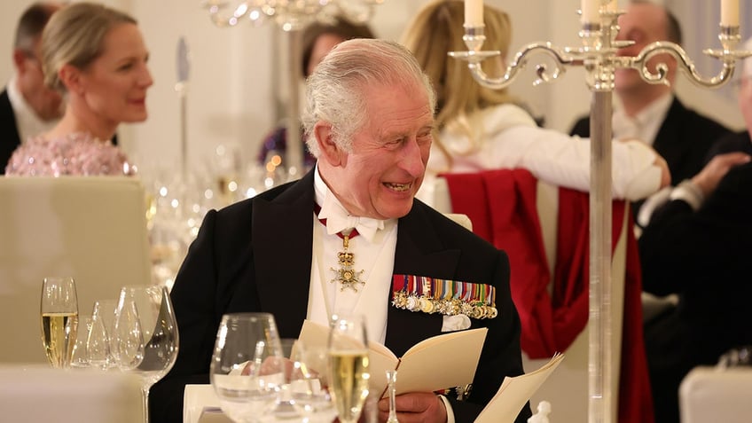 King Charles in a suit and medals smiling from a dining table.