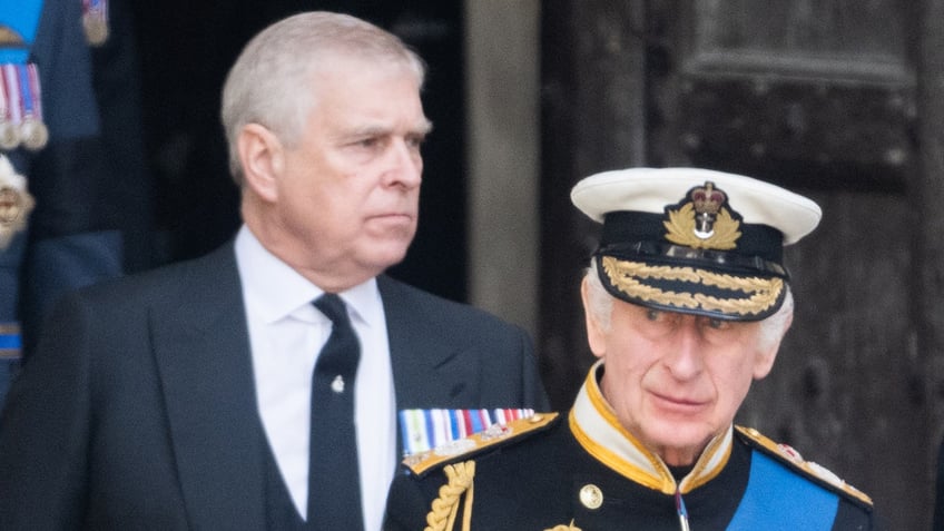 A close-up of King Charles in uniform walking ahead of Prince Andrew in a suit.