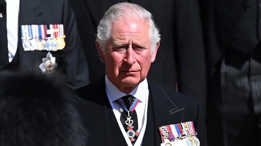A close-up of King Charles wearing a black suit and medals looking sad