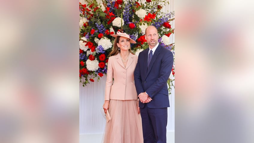 Kate Middleton wearing a pink Dior suit standing next to Prince William in a blue suit with flowers behind them.