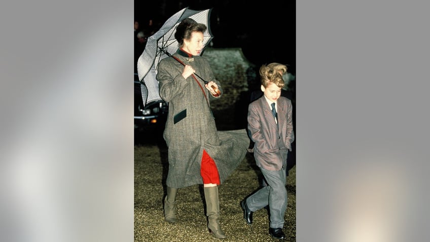 Princess Anne wearing a coat and holding an umbrella walking next to Prince William in a suit.