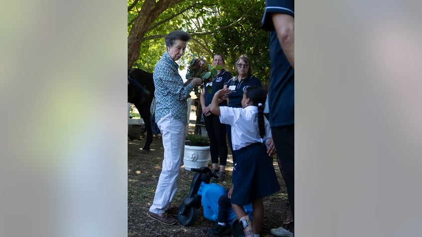 Princess Anne wearing a blue multiprinted blouse and white pants interacting with people outdoors.