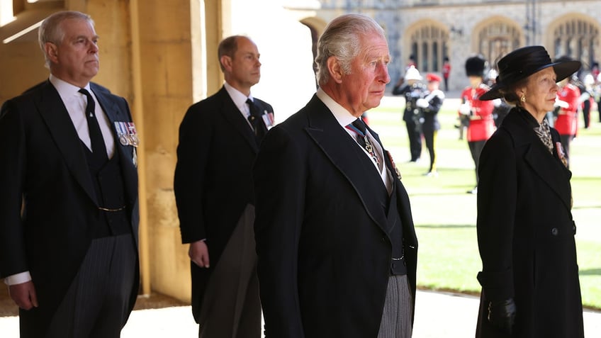 King Charles looking somber standing in front of Prince Andrew