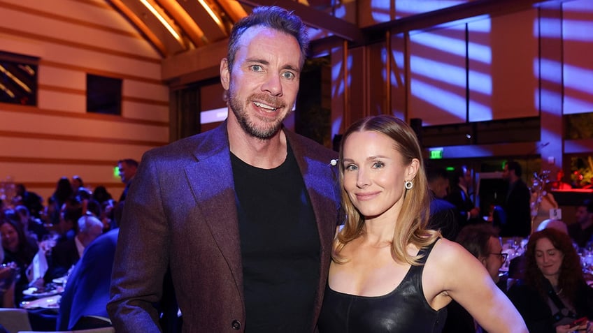 Dax Shepard smiles for a photo wearing a black shirt and purple jacket with wife Kristen Bell in a black dress who is soft smiling