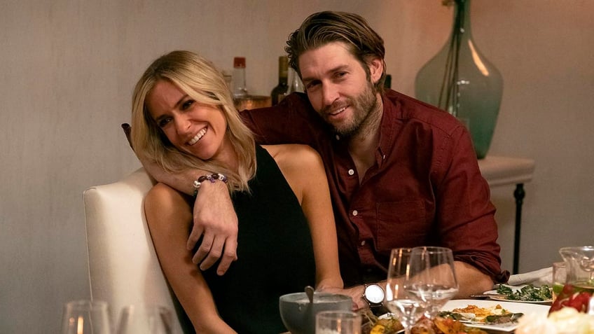 Kristin Cavallari in a black top leans as she sits at a table with Jay Cutler, who's wearing a maroon shirt and has his arm wrapped around Kristin