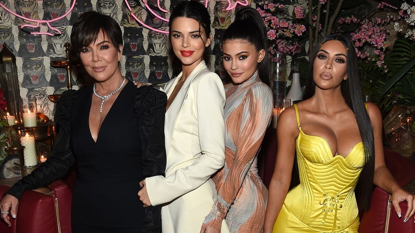 Kris Jenner in a black suit is held onto by daughter Kendall Jenner in a cream blazer and Kylie Jenner in a sparkly dress with Kim Kardashian in a yellow dress next to her