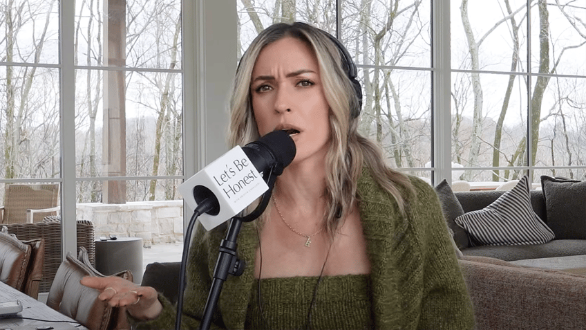 Kristin Cavallari in an olive green sweater set looks directly at the camera as she speaks on her podcast, wearing big black headphones