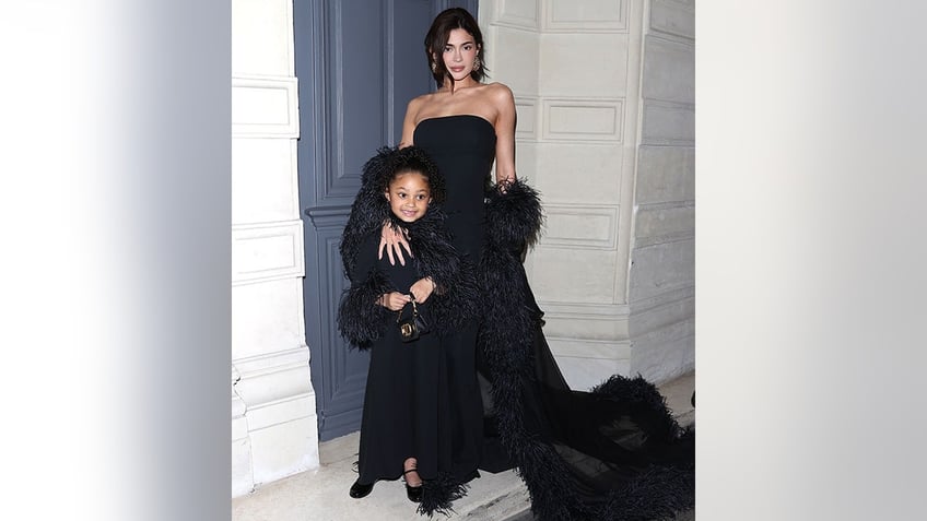 Kylie Jenner in a black strapless dress with a feather cape holds on to her daughter Stormi, also in black, in Paris while posing for a photo