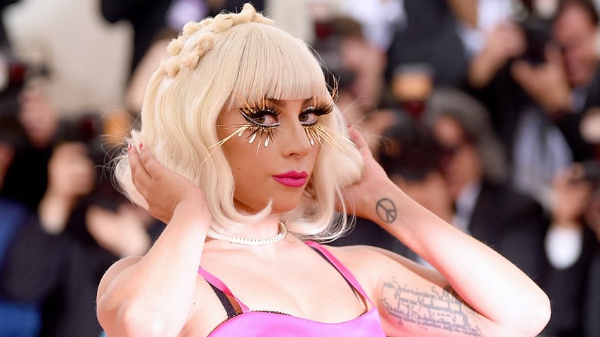 Lady Gaga plays with her short blond hair on the Met Gala red carpet, wearing a pink dress with extremely long eyelashes