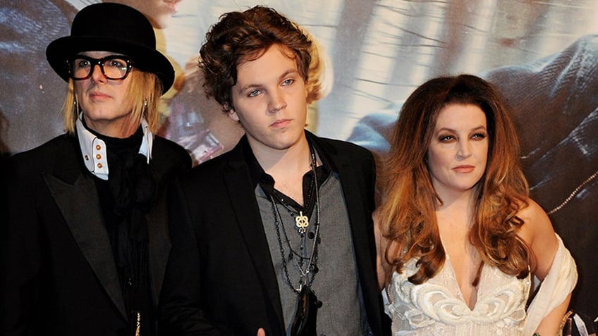 Michael Lockwood in a black jacket and hat is on the carpet with step-son Benjamin Keough in a black jacket standing next to mother Lisa Marie Presley in a white dress