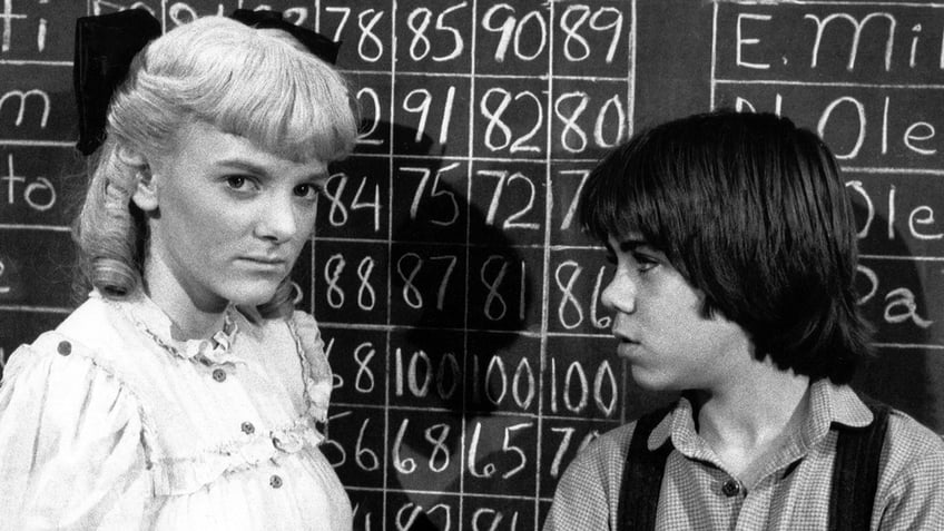 Alison Arngrim in costume looking stern in front of a chalkboard standing next to a little boy looking at her.