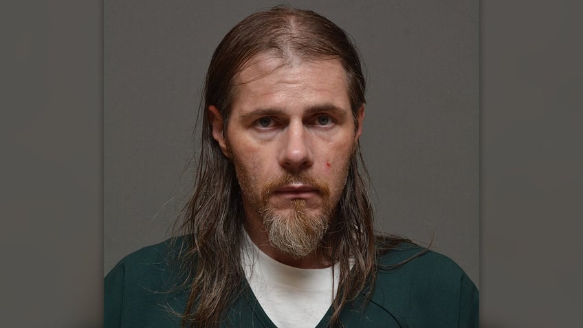 Ron Schroeder in green jail jumpsuit with a white udnershirt, long hair and graying goatee.