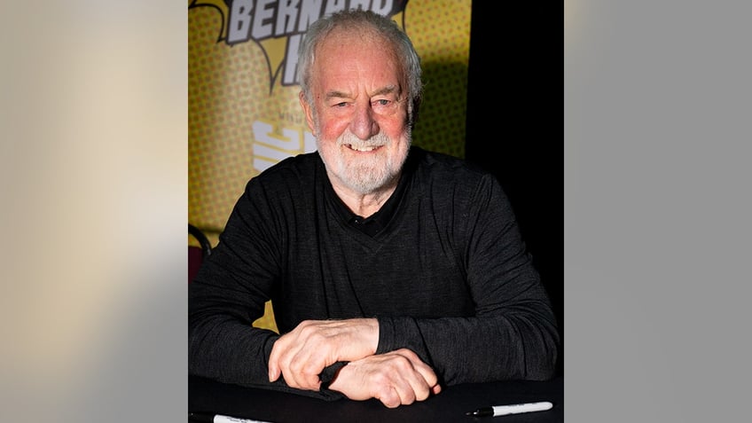 Bernard Hill smiles for a photo behind a table wearing a black shirt