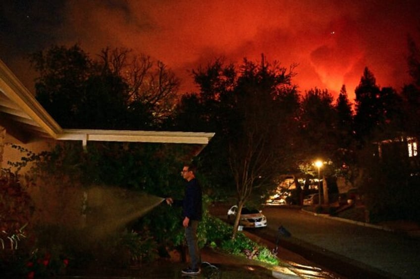 A man waters the front of his house as smoke and flames from the Palisades Fire burn towar