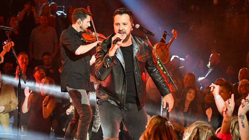 Luke Bryan in a leather jacket and black shirt performs onstage at the CMA Awards