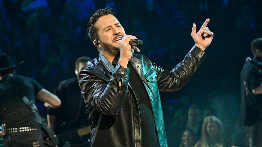Luke Bryan in a black leather jacket looks up and sings on stage