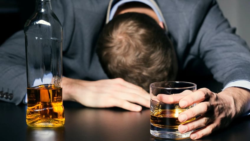 A man seen with head down on bar and hand around glass of liquor with bottle nearby.
