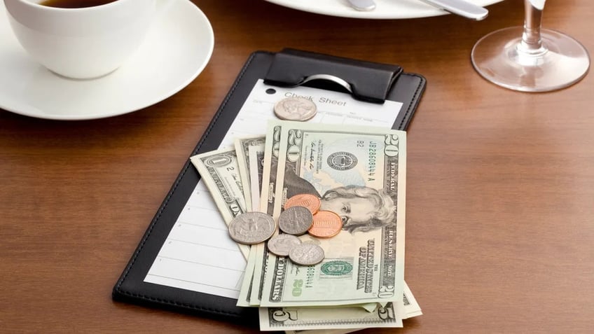 Cash and coins are seen on top of a restaurant check on a dinner table with coffee cup and wine glass seen in background.