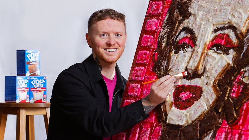 Man with portrait of Marilyn Monroe that is made out of pop tarts. He is smiling at the camera and has a paint brush on Marilyn's nose area.