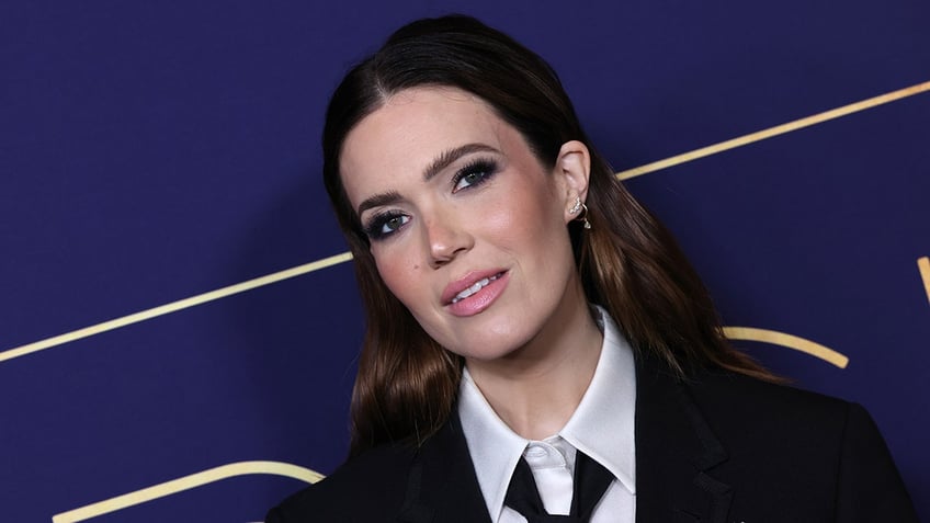 Mandy Moore in a black jacket, tie and white button up shirt looks at the camera