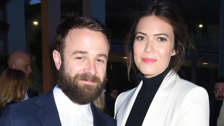 Taylor Goldsmith in a black suit and white shirt stands next to wife Mandy Moore in a white suit and black shirt