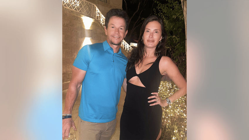Mark Wahlberg in a blue shirt smiles next to wife Rhea Durman in a black dress with a chest cut out with her hand on her hip