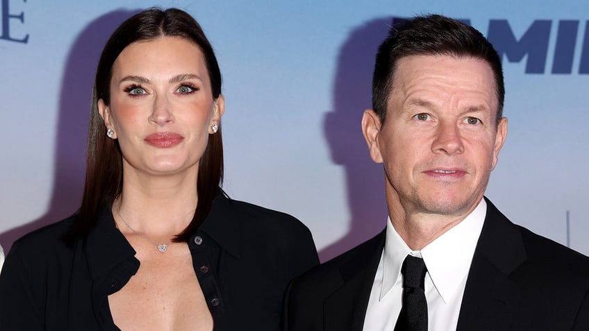 Rhea Durham in a black cardigan soft smiles on the carpet with husband Mark Wahlberg in a black suit and tie