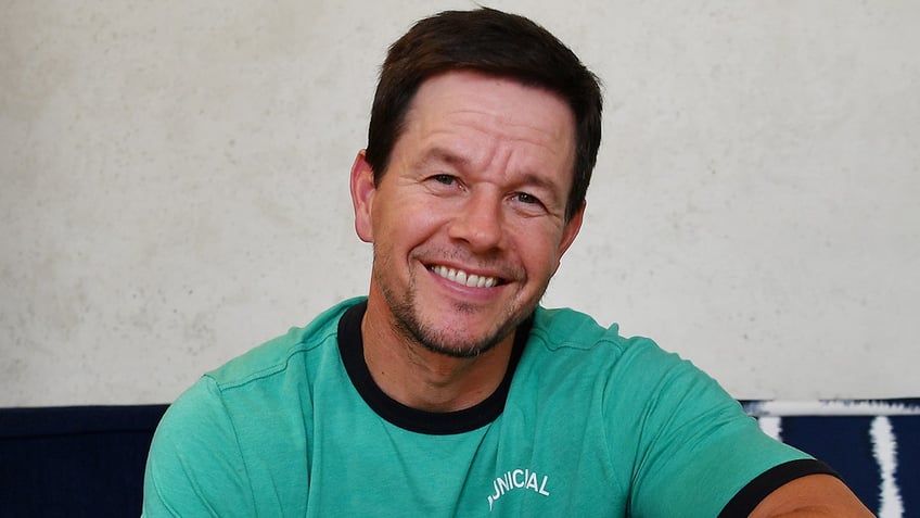 Mark Wahlberg poses in a teal t-shirt smiles for a picture why showcasing his tequila