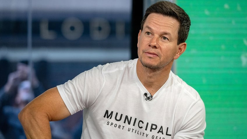 Mark Wahlberg in a white t-shirt outstreches his arm showing off his muscle