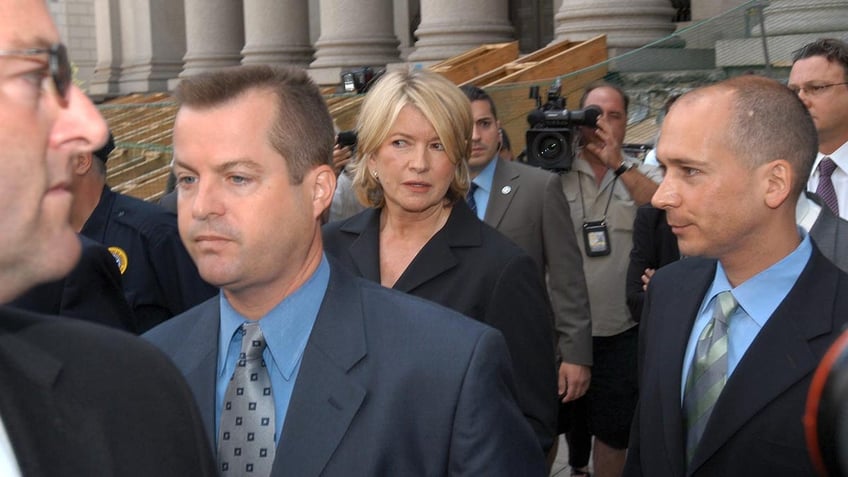 Martha Stewart in a black suit looking perplexed in a crowd of lawyers and reporters after being sentenced to prison