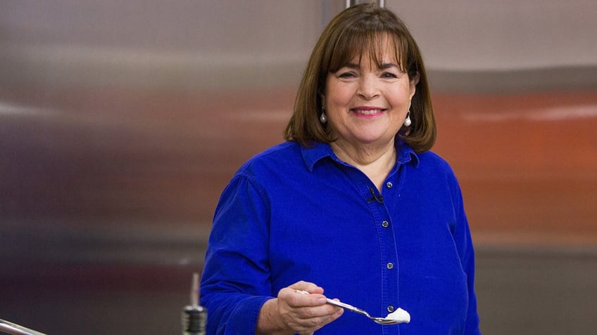 Ina Garten smiles in a bright blue shirt while holding a spoon up on camera