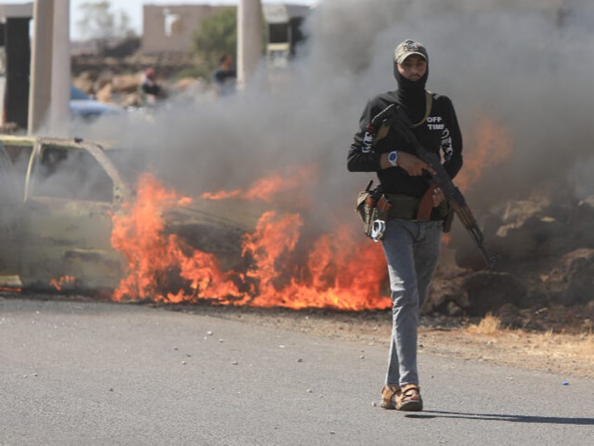 A member of the Syrian security forces walks past a burning car in the Mazraa area, near S