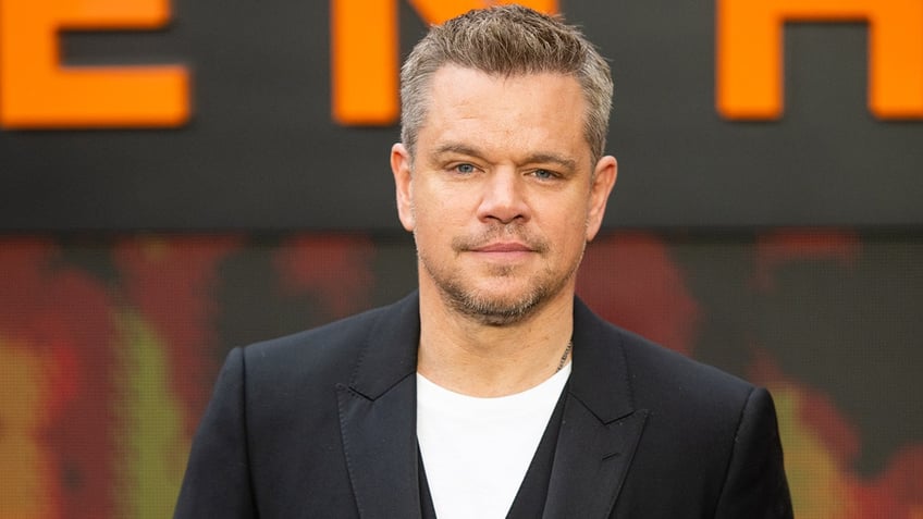 Matt Damon in a black suit stares directly at the camera on the carpet