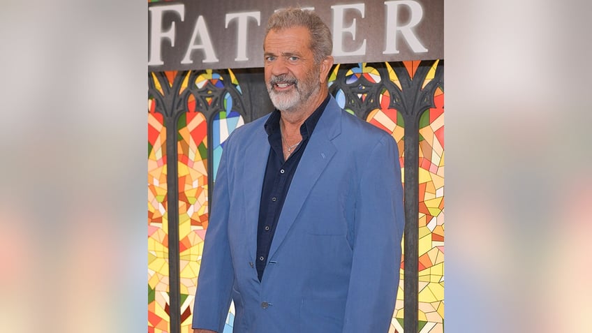 Mel Gibson smiles in a blue suit and black shirt on the carpet